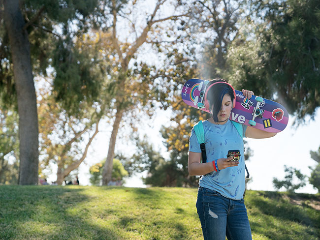 Teen outside holding a skateboard and looking at phone.