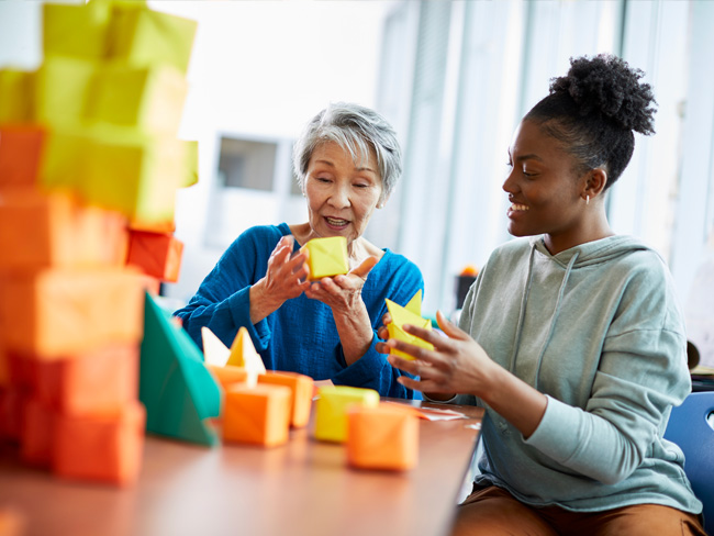 elderly woman seated at table with a younger Black woman doing crafts