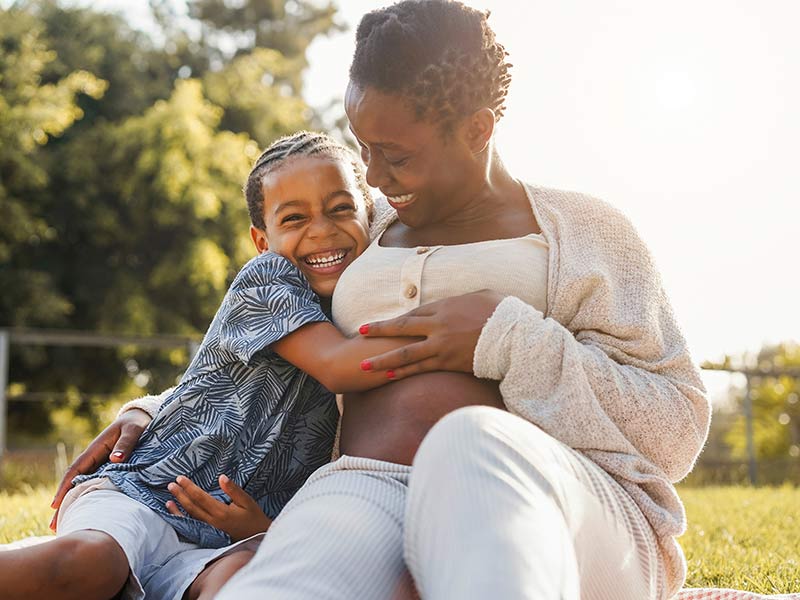 smiling pregnant mother with son hugging and sitting on grass at city park