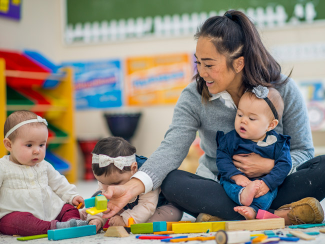 woman seated on floor with 3 babies playing with blocks