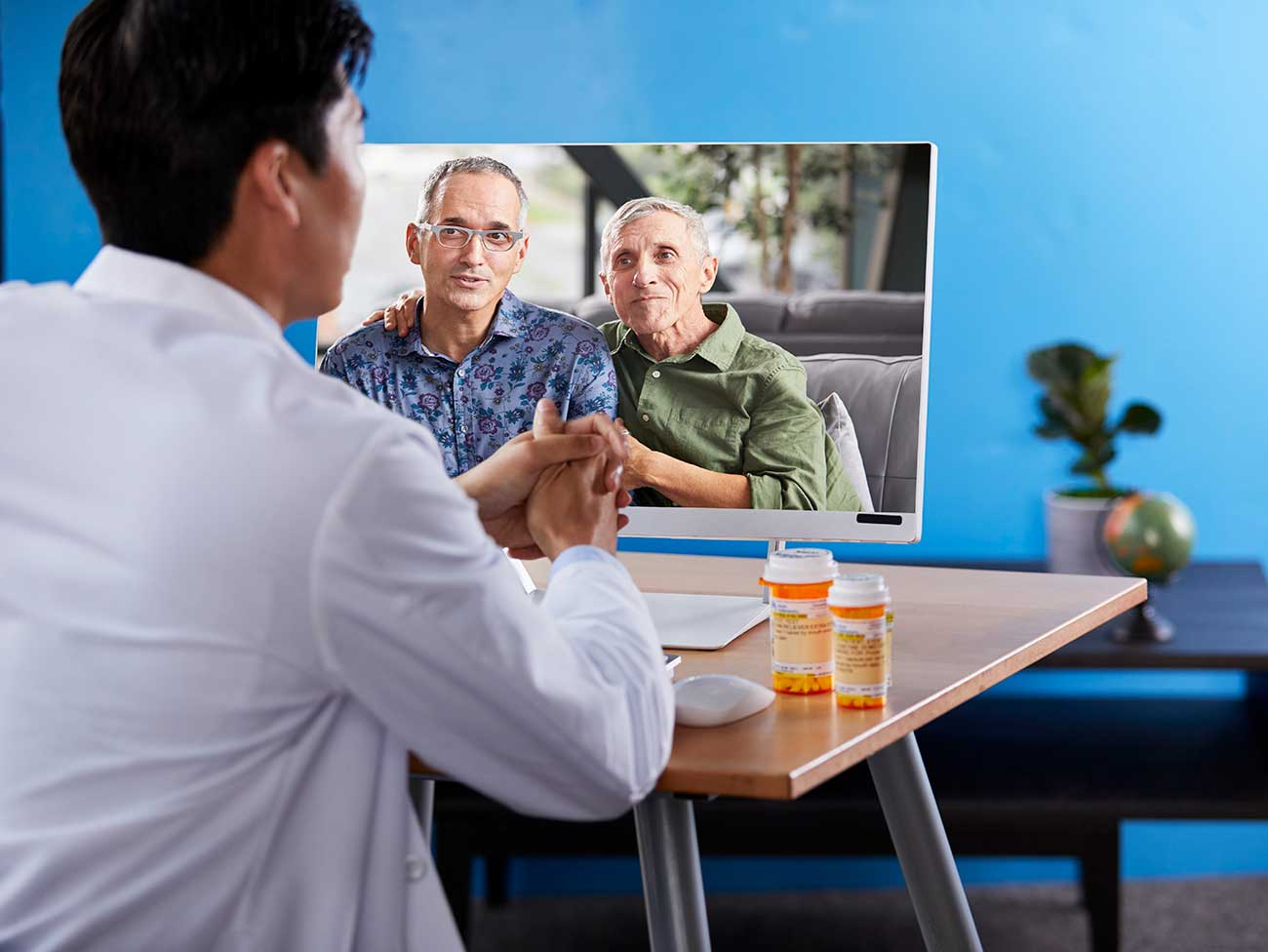 doctor wearing white coat seated at desk speaking to 2 older men through the computer