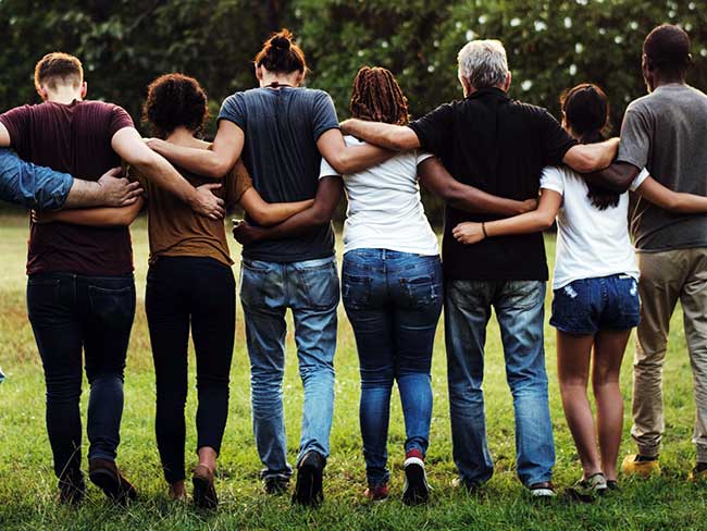 A group of friends, arms around each other's waists walking in a field as the sun rises. 