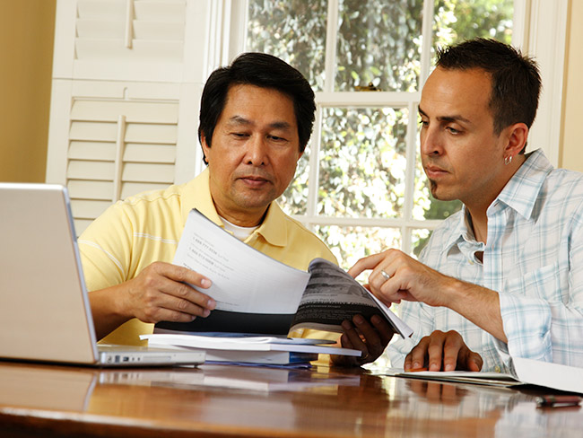 2 men seated at table with a laptop reviewing books and documents