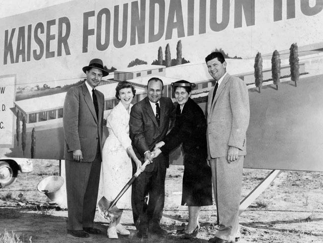 Five individuals, together holding a shovel, are standing in front of a large sign that reads: KAISER FOUNDATION HOSPITAL at the 1950 groundbreaking ceremony.