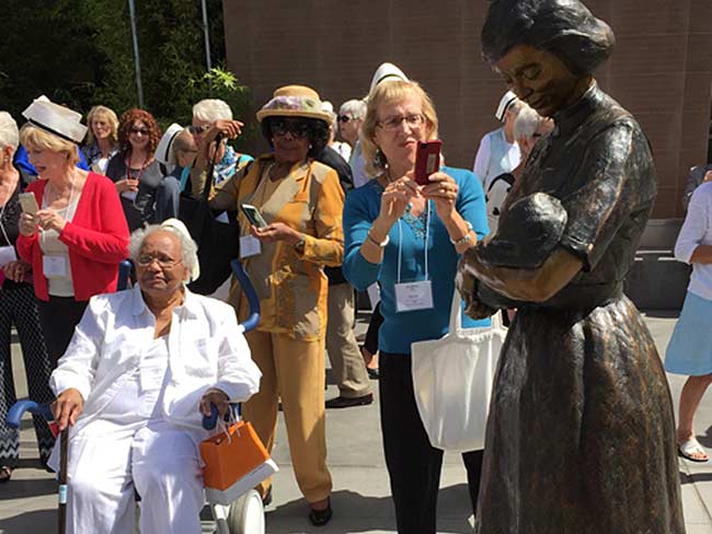 A crowd of women surround a sculpture of a curse cradling a baby in her arms.