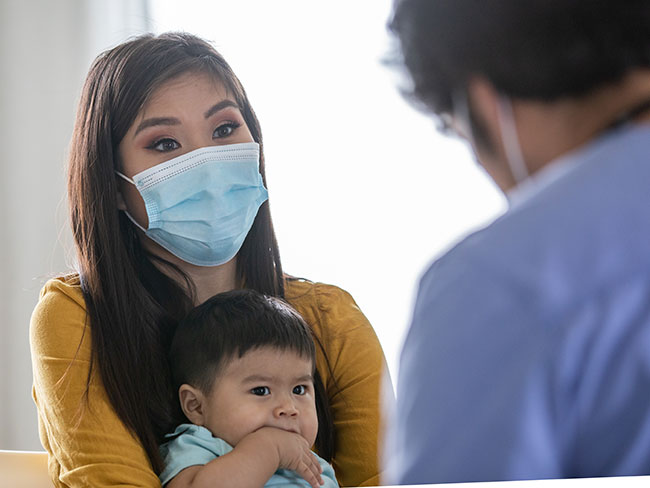 Mother in facemask with infant in lap during exam.