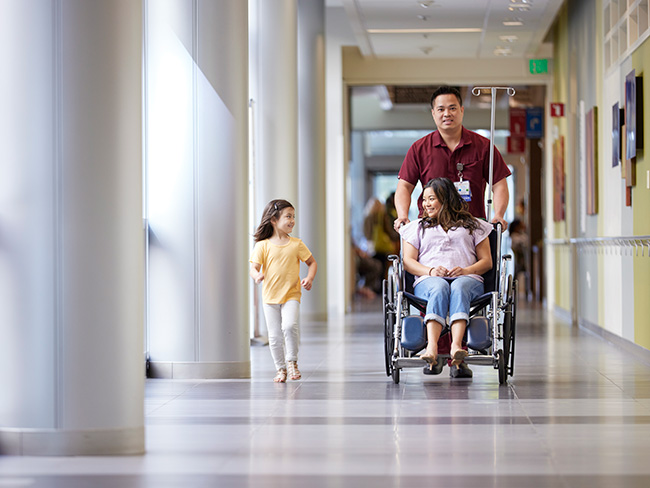 Nurse pushes mom in wheelchair with daughter walking nearby