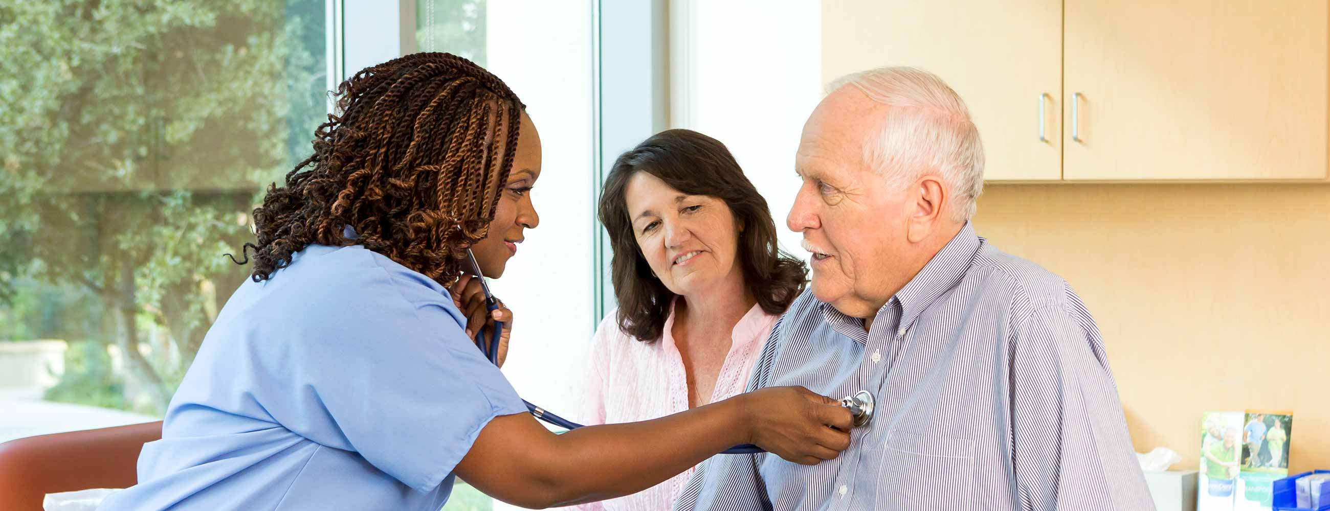 Senior man with his wife, in an exam room while a doctor holds a stethoscopes to his chest.