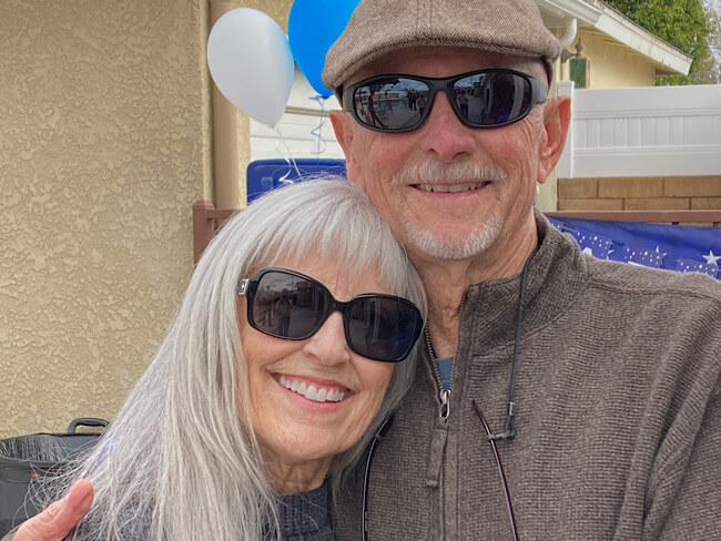 An older man smiles and puts his arm around the shoulder of an older woman.