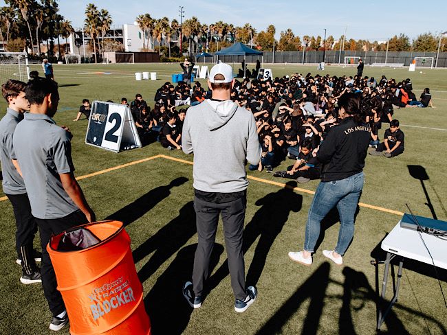 A few individuals stand at the front of a soccer field and speak to a large group of children who are wearing black shirts.