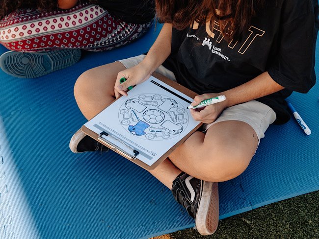 A young person colors a coloring page that includes a blue sports jersey and soccer balls.