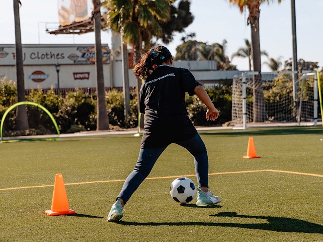 A girl kicks a soccer ball between two orange cones. 