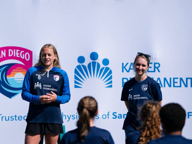 Two women wearing sports jerseys stand and speak to a group of children. 