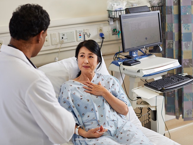 A woman rests in a hospital bed while she speaks to her physician, who stands near her.