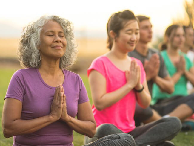 A group of women sitting cross-legged with palms meeting at their heart's center. Their eyes are closed, and they appear to be breathing deeply. 