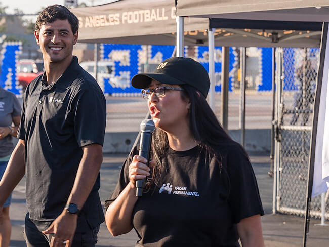 Carlos Vela stands next to Dr. Luna, who speaks into a microphone she is holding. They are both outside.