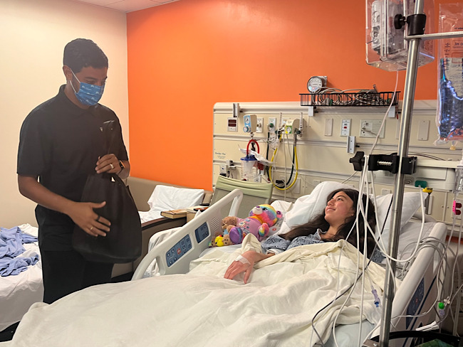 Carlos Vela, wearing a mask and holding a bag, stands next to the hospital bedside of a young girl who smiles at hi,m.