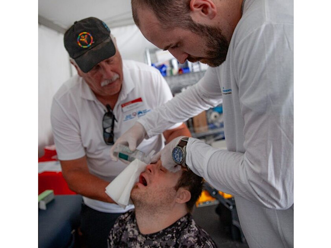 A man wearing gloves holds a cloth and a bottle filled with liquid to another man's eye. A third man stands nearby, assisting.