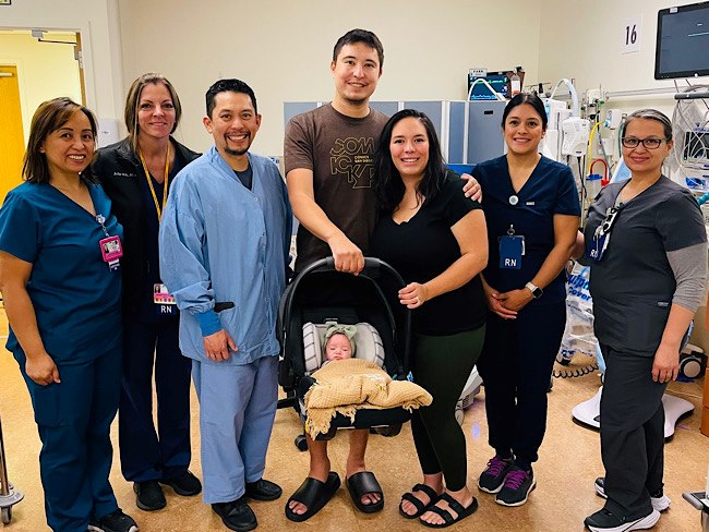 A man and woman hold a baby in a car seat. They are surrounded by medical professionals wearing scrubs.