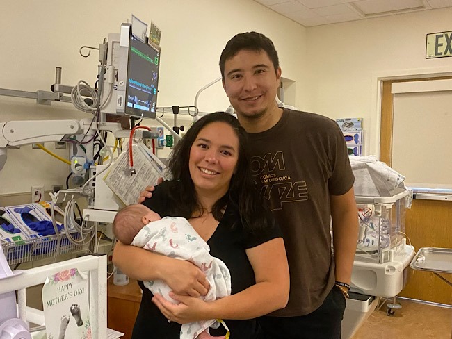 A man stands over a woman who is holding a baby. They are in a hospital, but they are smiling.