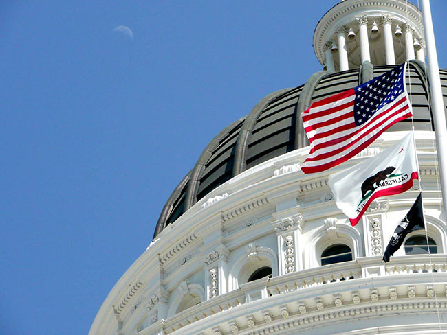 California Capitol building dome with the U.S. and California state flags flying in the wind in front of it.