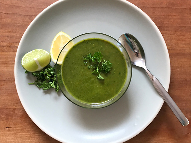 A bowl of green soup on a white plate with a spoon on the side. 