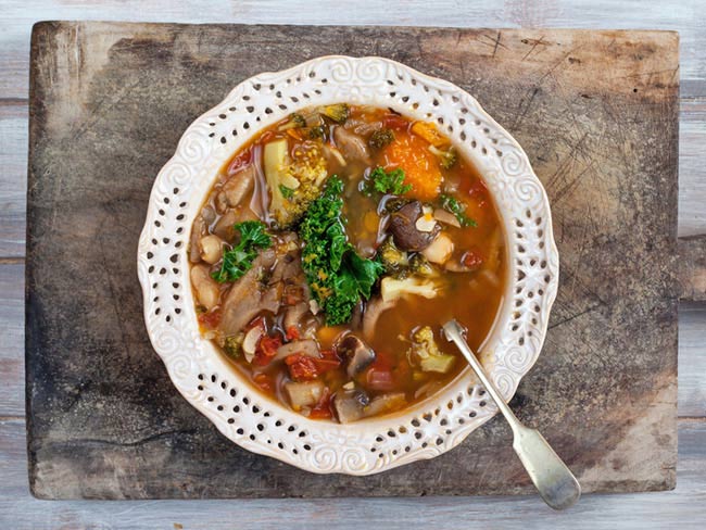 Kale and roasted vegetable soup in a fancy white bowl on a wooden cutting board.