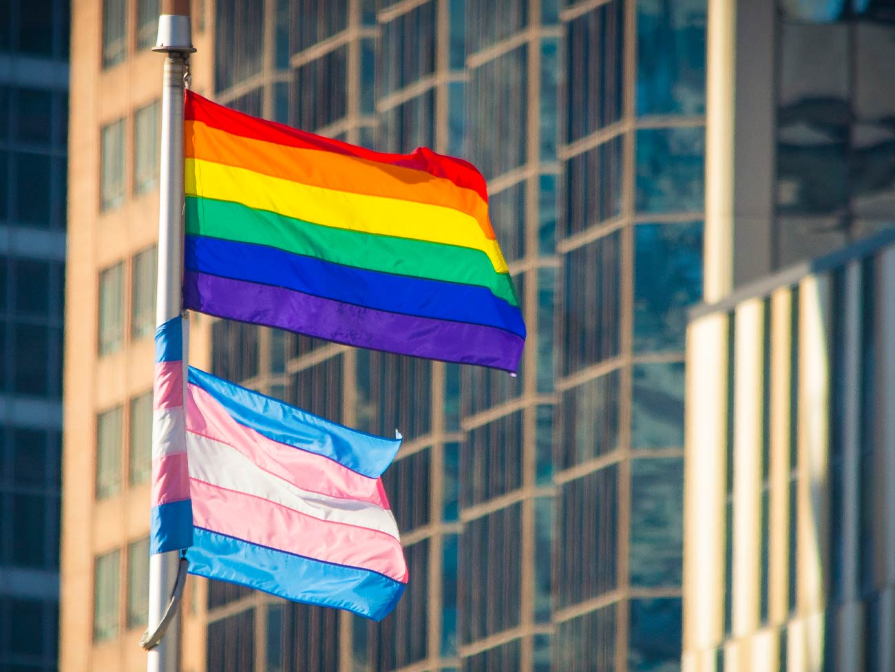 Pride and Trans Flag waving in the wind in a downtown urban setting. Office buildings can be seen behind.