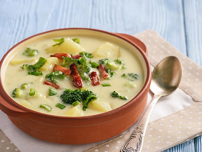 A crock filled with vegan potato broccoli soup with a spoon on the table to the right. 