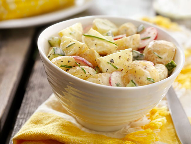 Creamy potato salad in white bowl on yellow napkin on wooden table.